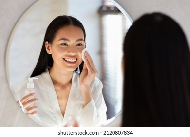 Attractive long-haired millennial asian lady in bathrobe using cleansing milk, removing makeup in front of mirror at bathroom, woman using facial tonner and cotton pad, over shoulder photo