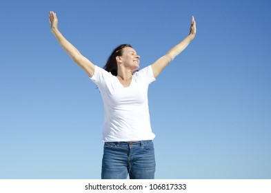 An Attractive Looking Mature Woman In Her Fifties Showing Her Happy Feelings By Rising Her Arms Up In The Air, With A Friendly Smile And Closed Eyes, Clear Blue Sky As Background And Copy Space.