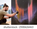 Attractive male artist is painting picture with paint spray can spraying it onto canvas at outdoor street exhibition, side view of man art maker