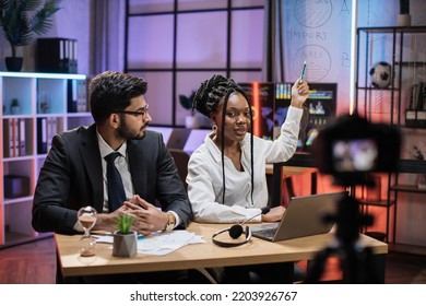 Attractive Male And Female Financiers, Arab Manager And Confident African American Business Woman Explaining Online Economic Charts On Glass Board To Their Colleagues Sitting In Front Of Video Camera.