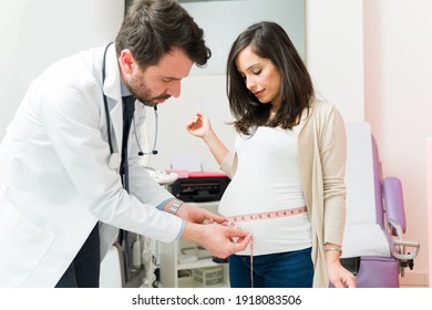 Attractive male gynecologist measuring the round belly of a beautiful pregnant woman at the examination room