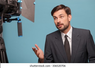 Attractive Male Tv Newscaster Is Reporting And Looking At The Camera. He Is Smiling And Gesturing. The Man Is Wearing A Suit. Isolated On Blue Background