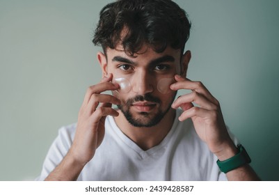 Attractive man applying eye patch facial with collagen looking at camera, isolated over grey background. Male morning daily beauty procedures. Skincare and
