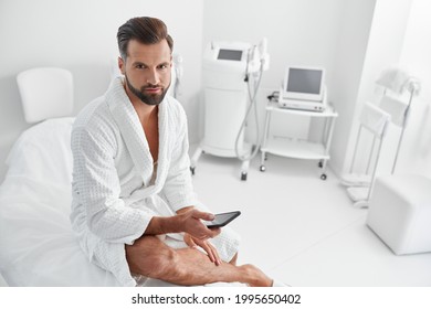 Attractive Man In Bathrobe With Smartphone Sits On Couch In Cosmetologycal Salon