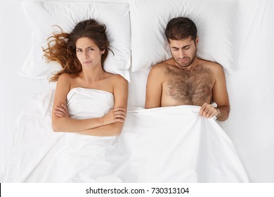 Attractive man feeling disappointed and depressed because of erectile dysfunction, looking at his penis under white cover, doesn't know what's the problem, angry wife lying next to him and waiting