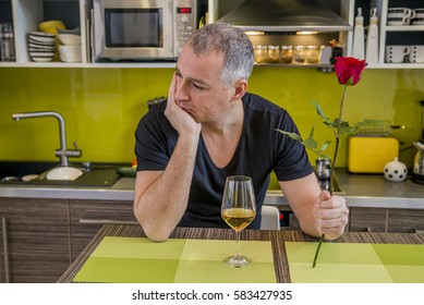 Attractive Man With Flowers In His Hand Is Waiting. The Thoughtful Man In Kitchen, Waits For The Woman. She Is Not Coming. Depressed Young Man Holding Single Rose