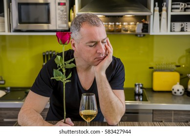 Attractive Man With Flowers In His Hand Is Waiting. The Thoughtful Man In Kitchen, Waits For The Woman. She Is Not Coming. Depressed Young Man Holding Single Rose