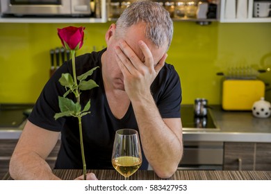 Attractive Man With Flowers In His Hand Is Waiting. The Thoughtful Man In Kitchen, Waits For The Woman. She Is Not Coming. Depressed Young Man Holding Single Rose