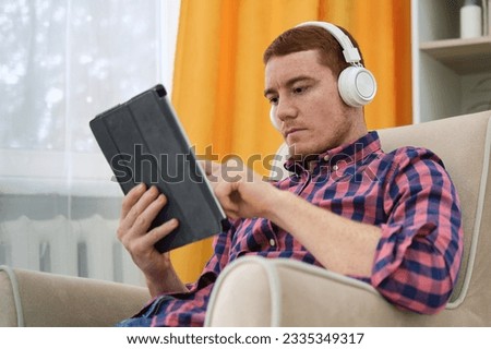 attractive man with freckles in a red plaid shirt with headphones and tablet listening to music at home, sitting in armchair in living room interior. Millennial man enjoying his favorite song