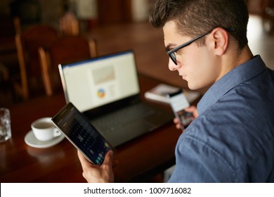 Attractive man in glasses working with multiple electronic internet devices. Freelancer businessman has laptop and smartphone in hands and laptop on table with charts on screen. Multitasking theme. - Shutterstock ID 1009716082