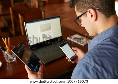 Attractive man in glasses working with multiple electronic internet devices. Freelancer businessman has laptop and smartphone in hands and laptop on table with charts on screen. Multitasking theme.