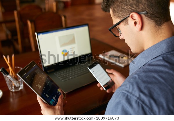 Attractive man in glasses working with multiple\
electronic internet devices. Freelancer businessman has laptop and\
smartphone in hands and laptop on table with charts on screen.\
Multitasking theme.