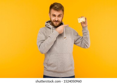 Attractive man in a gray hoodie points a finger at the credit card that is holding in his hand on a yellow background - image