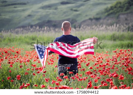 Attractive man holding Flag of the United States in beautiful poppy field on a clear, sunny day. Celebrating Independence Day, National holiday concept.