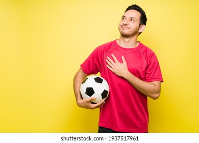 Attractive Man Holding A Soccer Ball Under His Arm And Touching His Chest With One Hand. Hispanic Man With A Red Sports Jersey Feeling Proud Of His Football Team 