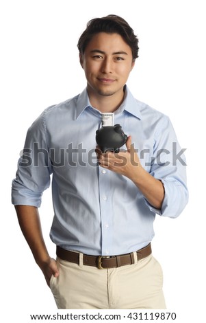 An attractive man in a light blue shirt and khaki pants standing against a white background smiling, holding a black piggy bank with a dollar bill sticking out.