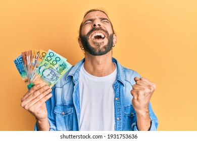 Attractive man with long hair and beard holding australian dollars screaming proud, celebrating victory and success very excited with raised arm 