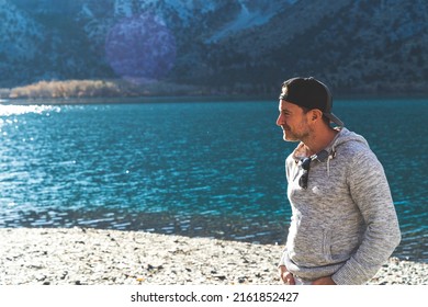 Attractive Man Standing In Front Of Mountain Lake With Ball Cap On Looking Away In Grey Hoodie.