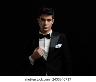 attractive man wearing black suit and bowtie fixing tuxedo collar while standing on black background
