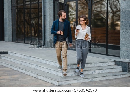 attractive man and woman going on stairs in urban city center in smart casual business style, talking, working together, smiling, stylish freelance people, holding coffee and laptop, discussing