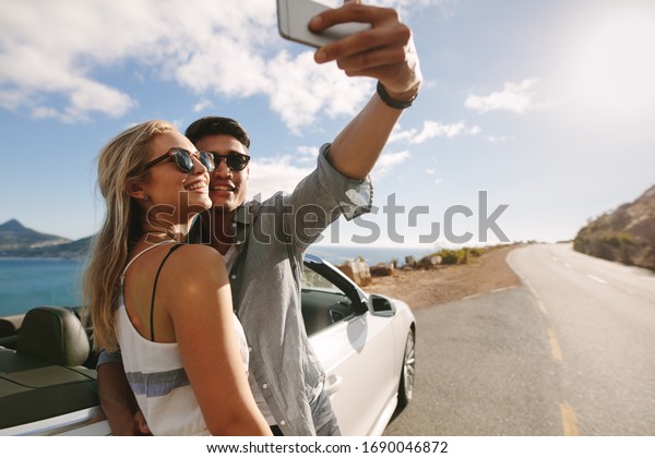 Attractive man and woman\
wearing sunglasses taking selfie photo together while standing by\
car on highway.