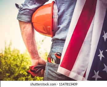 Attractive man in work clothes, holding tools and a US flag in his hands and looking into the distance against the background of trees, blue sky and sunset. View from the back. Labour Day Concept - Shutterstock ID 1147813409
