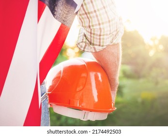 Attractive man in work clothes, holding American Flag and construction hard hat in his hands against green trees and blue sky. Rear view, closeup, outdoors. Labor and employment concept - Shutterstock ID 2174492503
