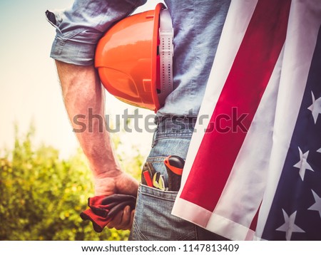 Attractive man in work clothes, holding tools and a US flag in his hands and looking into the distance against the background of trees, blue sky and sunset. View from the back. Labour Day Concept
