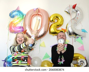 attractive man and young woman celebrating new year 2022 at home quarantine. boy holding alarm clocks. girl shooting confetti. big number balloons. silvester party. december 31. lockdown. january 1.