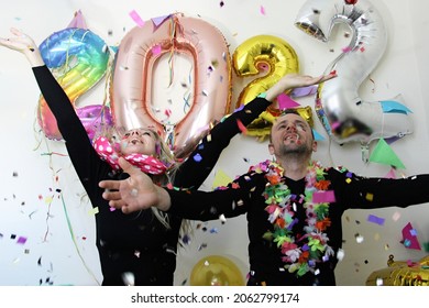 attractive man and young woman celebrating new year 2022 at home quarantine. boy and girl with hands up. big number balloons. silvester party. december 31. lockdown. confetti falling. january 1.
