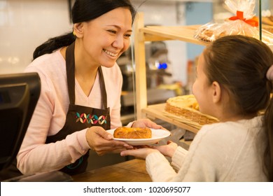 Attractive Mature Asian Female Baker Working At Her Bakery Store Smiling Joyfully Selling Pastries To A Little Girl Consumerism Profession Occupation Job Salesperson Small Business Owner Family