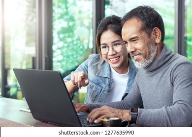 Attractive Mature Asian Man With White Stylish Short Beard Looking At Laptop Computer With Teenage Eye Glasses Hipster Woman In Cafe. Teaching Internet Online Or Wifi Technology In Older Man Concept.