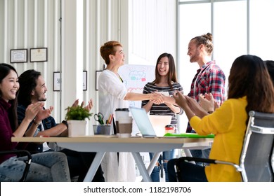 Attractive Mature Asian Woman Shaking Hand With Young Beard Hipster Caucasian With Welcome New Employee, Congratulating Successful Or Happy With Business Deal While Team Applauding Support Together.