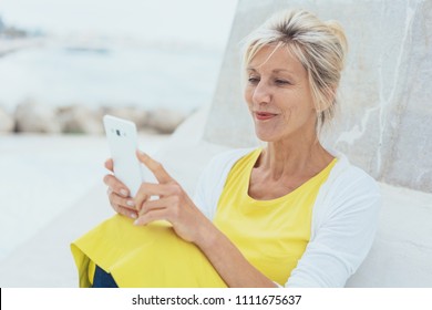 Attractive Mature Blond Woman Reading A Phone Message Seated On A White Bench In A High Key Portrait