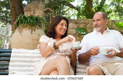 Attractive Mature Couple Sitting On A Bench In A Luxury Hotel Holiday Green Garden On Vacation, Drinking Coffee And Tea Together And Smiling Enjoying Retirement. Travel And Outdoors Senior Lifestyle.