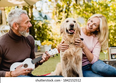 Attractive mature grey-haired man playing the ukulele for his beautiful blond wife and golden retriever in autumn park - Powered by Shutterstock