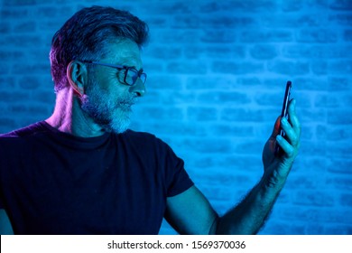 Attractive Mature Man In Eyeglasses Talking On Smartphone. Portrait Of Senior Man Talking On Phone. Portrait Of Man Stands Against A Brick Wall And Gel Light With Mobile Phone