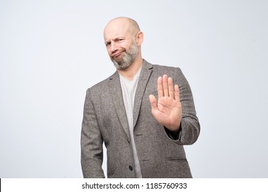 Attractive mature man showing refusal gesture. It is not for me, leave me in piece, has angry expression, poses against white concrete studio background