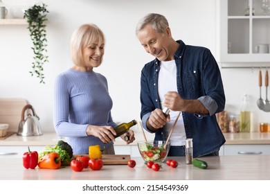 Attractive Mature Man And Woman In Casual Outfits Cooking Together At Home, Making Delicious Vegetable Salad, Adding Virgin Olive Oil, Seasoning, Kitchen Interior, Copy Space. Healthy Diet For Seniors