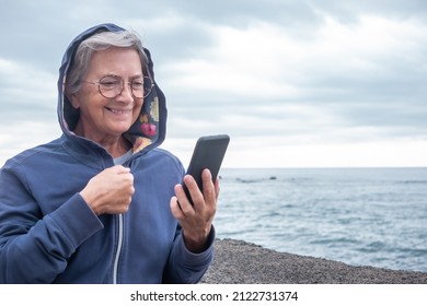 Attractive Mature Senior Pensioner Woman Enjoying Retirement At Sea. Caucasian Elderly Female Standing On The Beach In A Cloudy Day Using Mobile Phone Enjoying Tech And Social. Horizon Over Water