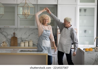 Attractive Mature Wife And Older Husband In Aprons Holding Hands Swirling Having Fun Distracted From Cooking In Fashionable Kitchen, Moving To Music, Dancing Together At Home Feel Carefree Look Happy