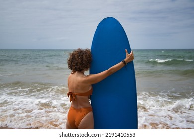 Attractive mature woman with curly hair, sunglasses and bikini, backside, posing holding a blue surfboard. Concept sea, sand, sun, beach, vacation, surf, summer.