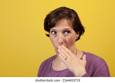 Attractive Mature Woman In Purple Blouse On A Yellow Background With Hand Over Mouth Looking Away To Camera Right