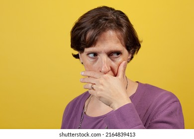 Attractive Mature Woman In Purple Blouse On A Yellow Background With Hand Over Mouth Looking Away To Camera Right