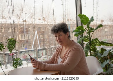 An Attractive Mature Woman Of Retirement Age Holds Her Smartphone In Her Hands In A Cafe Against The Background Of A Window. Online Dating