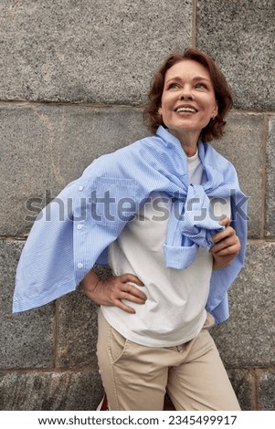 Attractive mature woman standing in a relaxed pose against the background of a stone wall. A middle-aged brunette in casual clothes looking up and smiling dreamily. Active lifestyle for elderly.