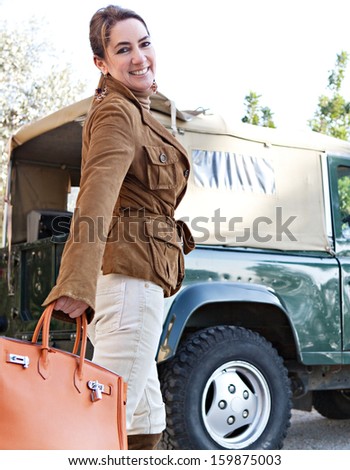 Attractive mature woman tourist getting ready for adventure while visiting a safari park on vacation with a four wheel drive car, turning to camera smiling during a sunny day outdoors.