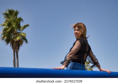 Attractive Mature Woman In Transparent Black Shirt And Jeans, Leaning On A Railing In Sensual And Provocative Attitude. Concept Maturity, Beauty, Fashion, Trend, Sensuality, Provocation.