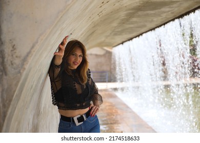 Attractive Mature Woman, With Transparent Black Shirt And Jeans, Posing Next To A Waterfall In A Sensual And Provocative Attitude. Concept Maturity, Beauty, Fashion, Seduction, Provocation.