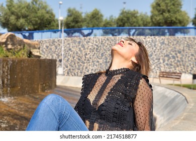 Attractive Mature Woman, Wearing Transparent Black Shirt And Jeans, Sitting Next To A Fountain, Posing In Sensual And Provocative Attitude. Concept Maturity, Beauty, Fashion, Seduction, Provocation.